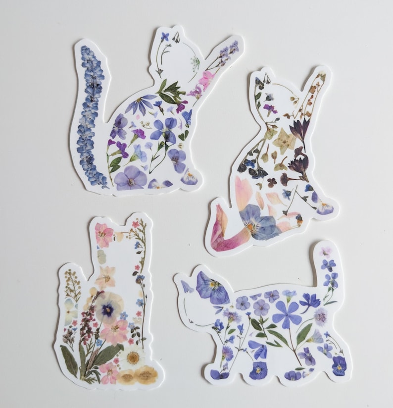 Puede incluir: Cuatro pegatinas con forma de gato hechas de flores prensadas. Los gatos est&aacute;n en varias poses y est&aacute;n decorados con flores moradas, azules, rosas y blancas.