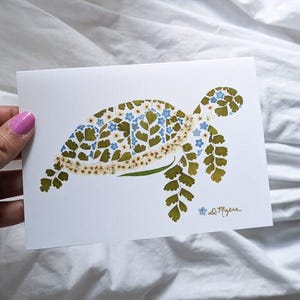 May include: A white card with a green and blue pressed flower design of a turtle. The artist's signature, "D. Myers", is in the bottom right corner.