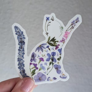 Puede incluir: Una pegatina blanca con una silueta de gato hecha de flores prensadas. Las flores son principalmente de tonos morados y azules, con algunas flores rosas.