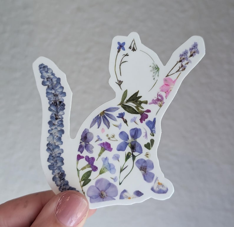 Puede incluir: Una pegatina blanca con una silueta de gato hecha de flores prensadas. Las flores son principalmente de tonos morados y azules, con algunas flores rosas.