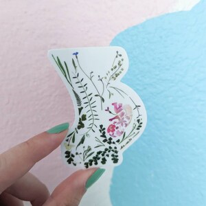 May include: A white sticker featuring a floral design in the shape of a pregnant belly. The flowers are pressed and dried, and include pink, white, and green colours.
