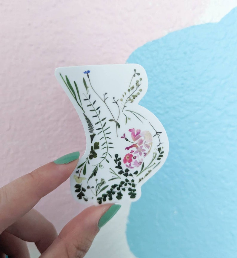 May include: A white sticker featuring a floral design in the shape of a pregnant belly. The flowers are pressed and dried, and include pink, white, and green colours.