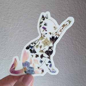 Puede incluir: Una pegatina de silueta de gato blanco hecha con flores prensadas. El gato est&aacute; sentado con la pata levantada. Las flores son de color rosa, azul, morado y verde.