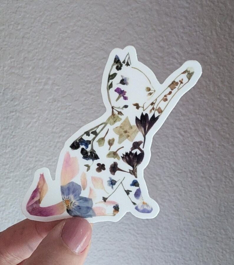 Puede incluir: Una pegatina de silueta de gato blanco hecha con flores prensadas. El gato est&aacute; sentado con la pata levantada. Las flores son de color rosa, azul, morado y verde.