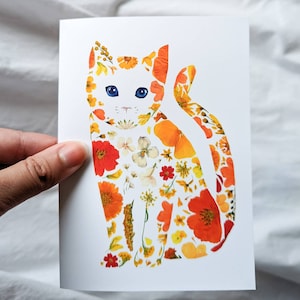 May include: A white greeting card featuring a cat silhouette filled with colorful pressed flowers. The cat has blue eyes and is composed of orange, yellow, and white floral elements. The card is held against a white fabric background.