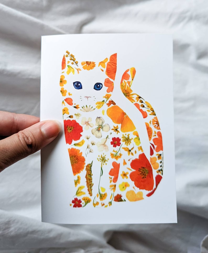 May include: A white greeting card featuring a cat silhouette filled with colorful pressed flowers. The cat has blue eyes and is composed of orange, yellow, and white floral elements. The card is held against a white fabric background.