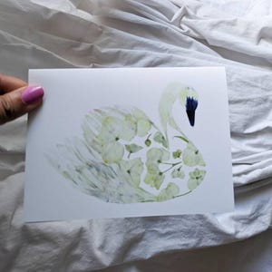May include: A white greeting card featuring a swan illustration. The swan is composed of light green and white floral elements, with a dark blue beak. The card is held against a white fabric background.