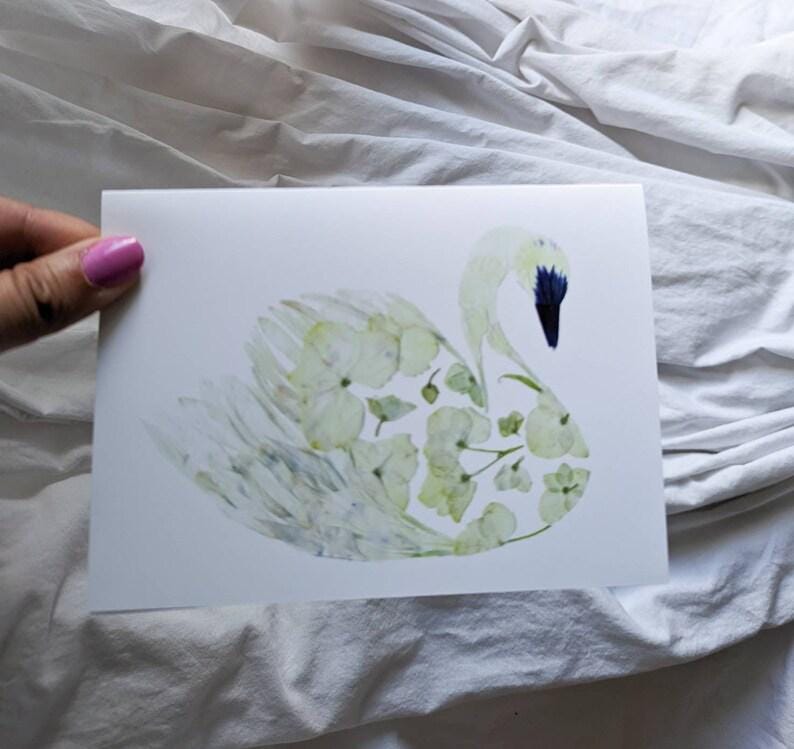 May include: A white greeting card featuring a swan illustration. The swan is composed of light green and white floral elements, with a dark blue beak. The card is held against a white fabric background.
