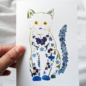 May include: A white card featuring a cat illustration adorned with blue and purple pressed flowers. The cat has yellow eyes and a long tail. The image is held by a hand.