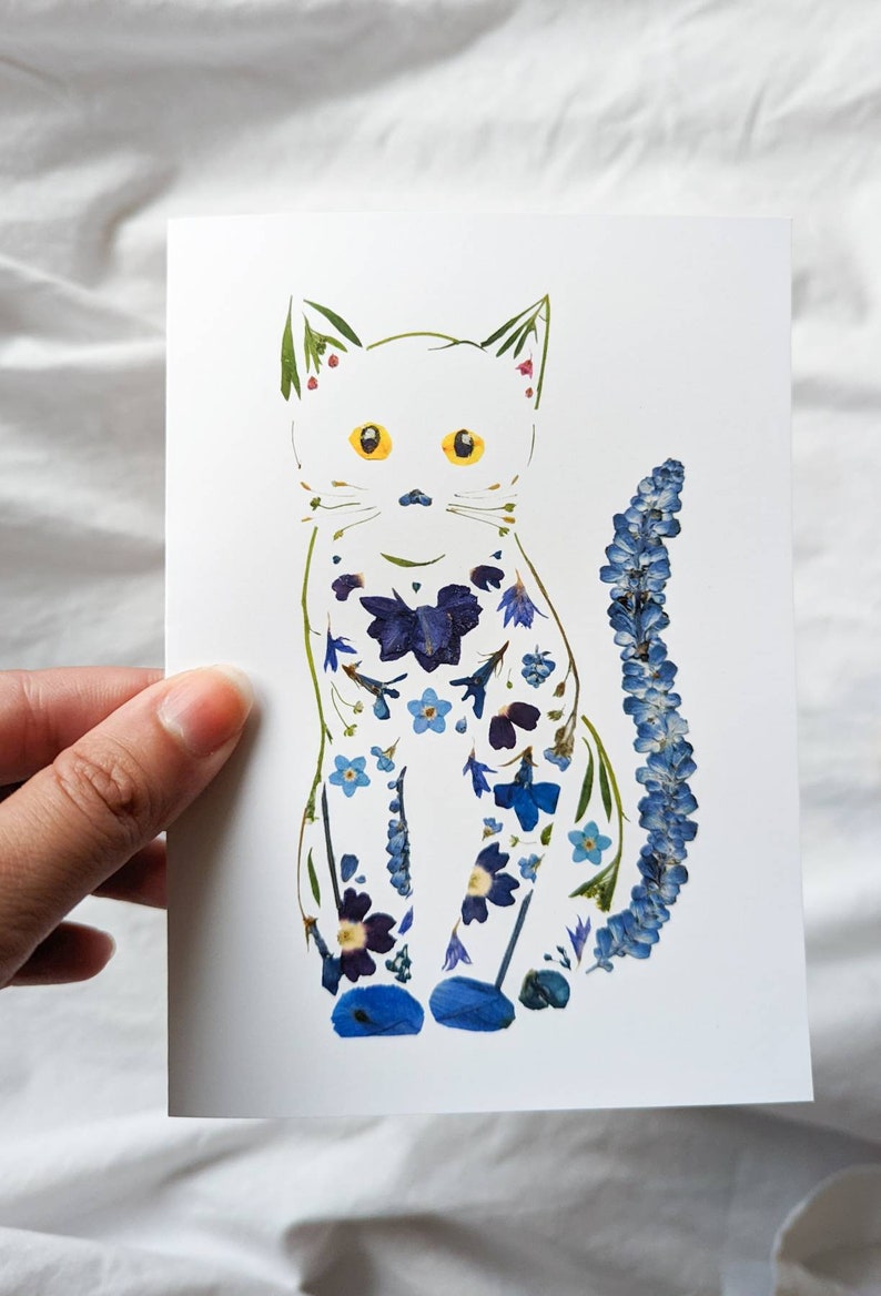 May include: A white card featuring a cat illustration adorned with blue and purple pressed flowers. The cat has yellow eyes and a long tail. The image is held by a hand.