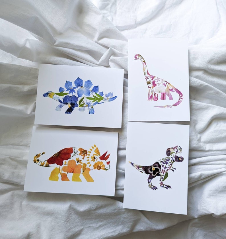 May include: Four cards with different dinosaurs made of pressed flowers. The dinosaurs are a stegosaurus, a brachiosaurus, a triceratops, and a tyrannosaurus rex. The cards are white with a black border.