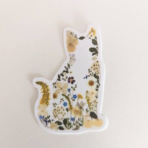 May include: A white cat silhouette sticker with pressed flowers and leaves in shades of yellow, white, blue, and green.
