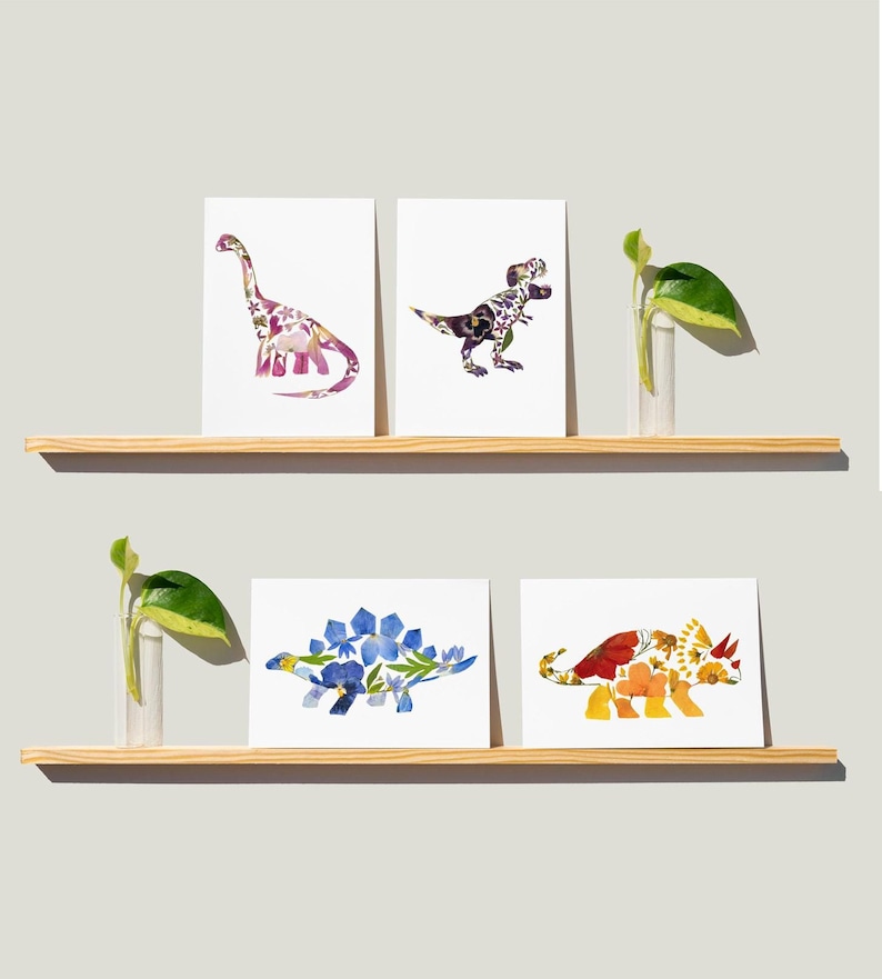 May include: Four framed prints of dinosaurs made from pressed flowers. The dinosaurs are a stegosaurus, a brachiosaurus, a tyrannosaurus rex, and a triceratops. The prints are on a white background and are displayed on a wooden shelf.
