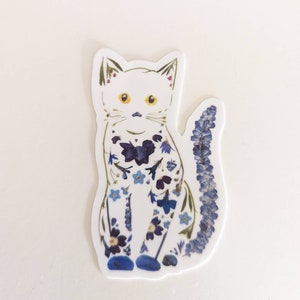 May include: A white cat sticker with a blue and purple floral design. The cat is sitting with its tail curled behind it.