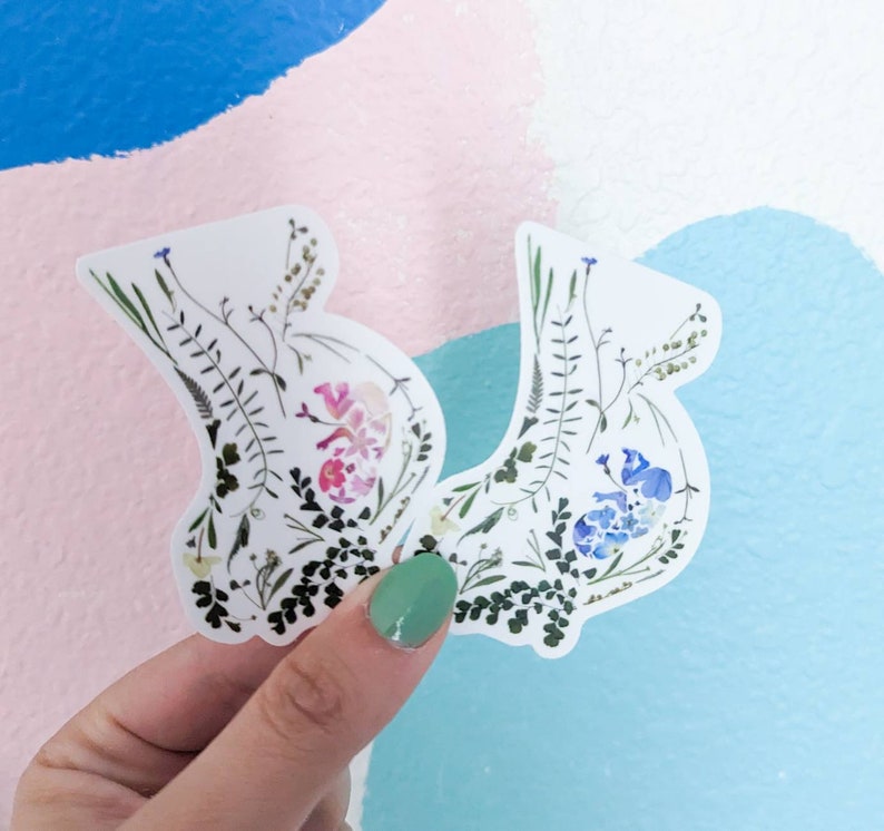 May include: Two white stickers with pressed flowers and leaves. One sticker features pink flowers and the other features blue flowers.
