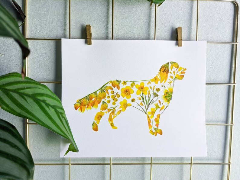 May include: A white print of a dog silhouette filled with yellow flowers and greenery. The artwork is clipped to a gold wire grid with a wooden clip. A green plant with striped leaves is in the foreground.