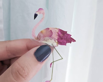May include: A pink flamingo sticker made from pressed flowers. The flamingo is standing on one leg with its head turned to the side. The sticker is clear and has a white background.