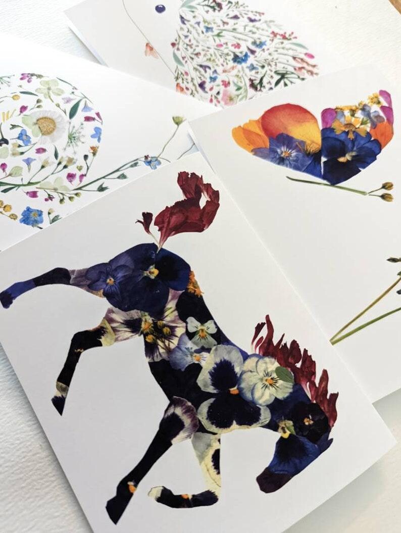 May include: A set of four cards featuring pressed flower art. Each card depicts a different design: a spiral, a butterfly, a horse, and a bird. The designs are made with a variety of colorful flowers.