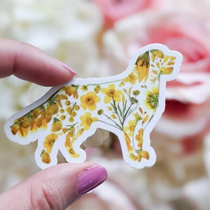 Puede incluir: Una pegatina blanca con un dise&ntilde;o floral amarillo en forma de perro. La pegatina est&aacute; siendo sostenida por una mano.