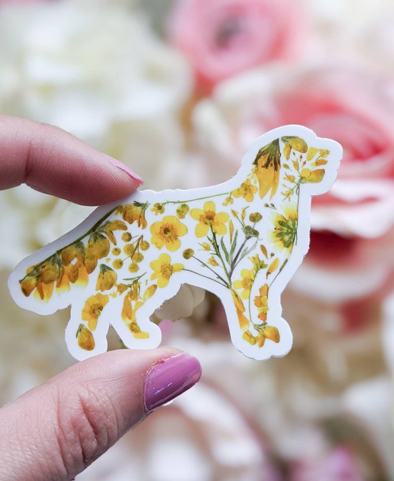 Puede incluir: Una pegatina blanca con un dise&ntilde;o floral amarillo en forma de perro. La pegatina est&aacute; siendo sostenida por una mano.