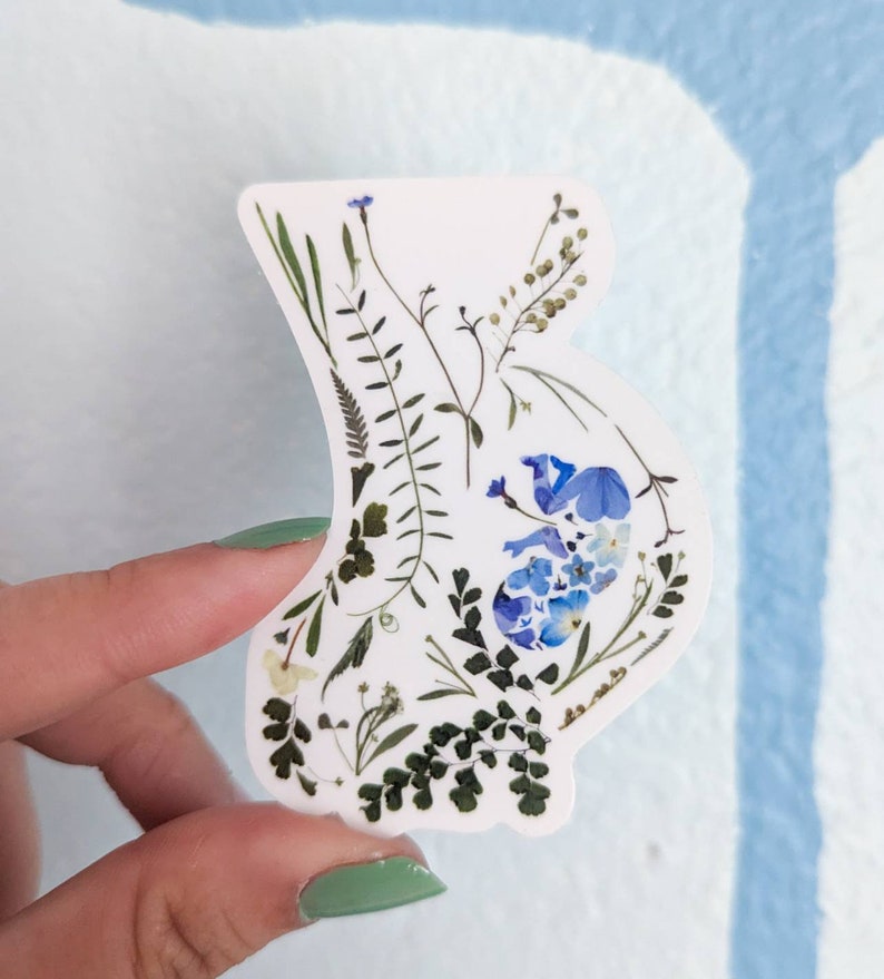 May include: A white sticker with a blue and green pressed flower design in the shape of a pregnant belly.