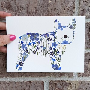 May include: A white card featuring a dog silhouette crafted from various blue pressed flowers. The floral arrangement includes different shades of blue blooms and green stems, creating a unique botanical artwork. The card is held against a brick wall.