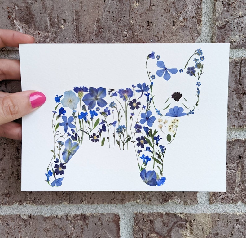May include: A white card featuring a dog silhouette crafted from various blue pressed flowers. The floral arrangement includes different shades of blue blooms and green stems, creating a unique botanical artwork. The card is held against a brick wall.