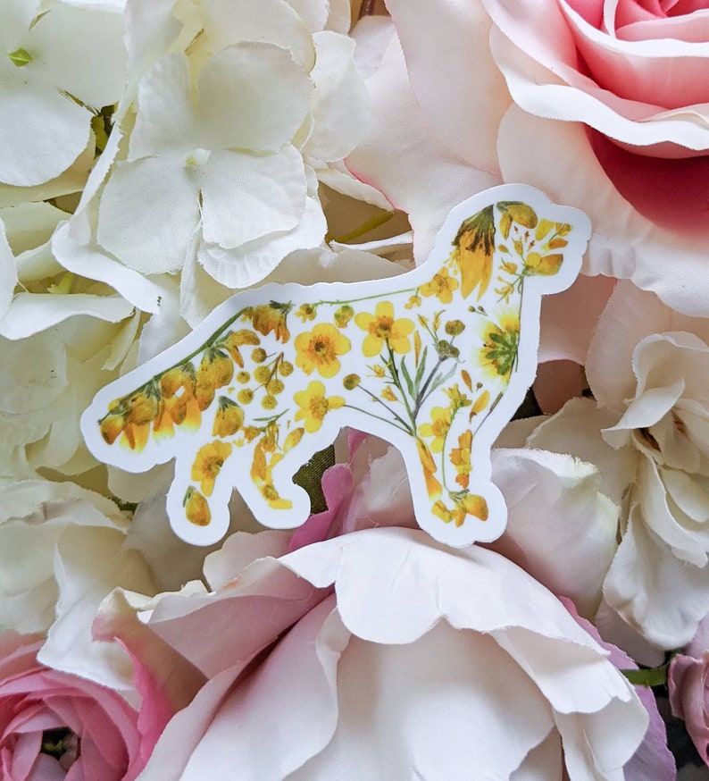 Puede incluir: Una pegatina blanca con una silueta de perro hecha de flores amarillas. La pegatina est&aacute; sobre un fondo de flores blancas y rosas.