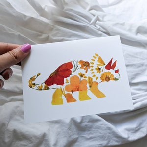 May include: A white card with a pressed flower art piece of a triceratops. The triceratops is made of pressed flowers in shades of orange, yellow, and red.