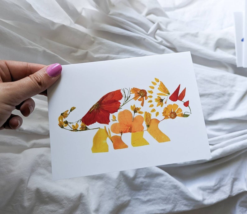 May include: A white card with a pressed flower art piece of a triceratops. The triceratops is made of pressed flowers in shades of orange, yellow, and red.