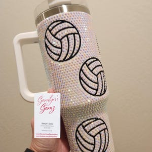 Puede incluir: Un vaso de acero inoxidable blanco con una tapa blanca y un asa. El vaso est&aacute; cubierto de diamantes de imitaci&oacute;n iridiscentes y presenta tres dise&ntilde;os de voleibol en blanco y negro. El vaso est&aacute; siendo sostenido por una mano.
