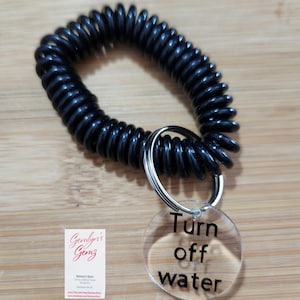 May include: Black coiled wrist keychain with a silver key ring and a clear acrylic tag. The tag has the words "Turn off water" in black. The keychain is on a wooden surface.