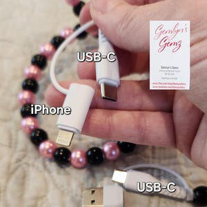 May include: White USB charging cable with multiple connector types: USB-C, iPhone, and USB-A. The cable is held up against a beaded bracelet with pink and black beads. The image also includes a business card with the text "Gemlyn's Gemz".
