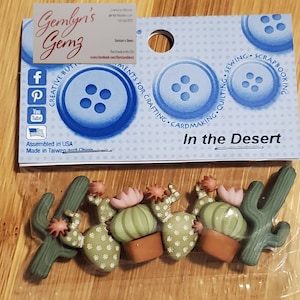 May include: A set of six green and brown cactus-themed buttons. The buttons are in the shape of cacti and have a white flower on top. The buttons are packaged in a clear plastic bag with a blue and white label that says "Gemlyn's Gemz" and "In the Desert".