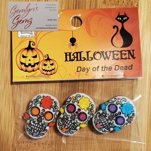 May include: Three colorful Day of the Dead sugar skull buttons with floral designs. The buttons are packaged on a card with a black cat and pumpkins.