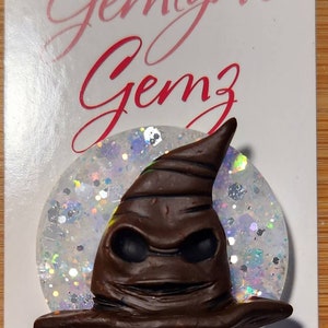 May include: A brown and black Sorting Hat from the Harry Potter series, with a white glitter background. The hat is a handmade resin charm. The text "Gemlyn's Gemz" is written on the card below the charm. The text "www.Etsy.com/shop/GemlynsGemz" and "www.facebook.com/GemlynsGemz" is written below the text.