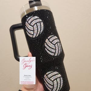 Puede incluir: Un vaso de acero inoxidable negro con un dise&ntilde;o de voleibol en blanco y negro. El vaso tiene un asa y una tapa. El vaso est&aacute; cubierto de diamantes de imitaci&oacute;n.