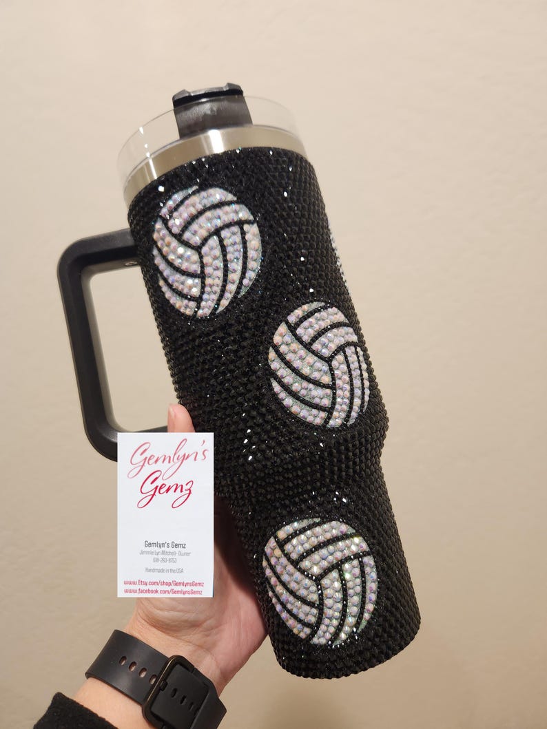 Puede incluir: Un vaso de acero inoxidable negro con un dise&ntilde;o de voleibol en blanco y negro. El vaso tiene un asa y una tapa. El vaso est&aacute; cubierto de diamantes de imitaci&oacute;n.