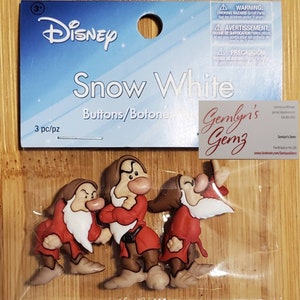 Könnte beinhalten: Drei Plastikknöpfe mit den sieben Zwergen aus Disneys Schneewittchen. Die Knöpfe sind rot, braun und weiß. Die Knöpfe sind mit "Gemly's Gemz" und "Handmade in the USA" beschriftet.