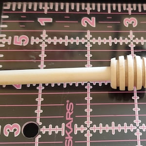 May include: A wooden honey dipper with a rounded handle and a grooved, spoon-shaped end. The dipper is approximately 5 inches long and is made of natural wood.