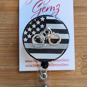 May include: A black and white American flag with a thin blue line and silver handcuffs on a round badge reel. The badge reel is attached to a clear plastic badge holder with a black retractable cord.