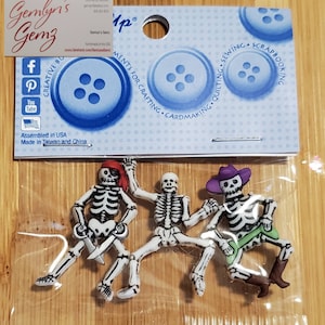 Puede incluir: Tres botones de pirata esqueleto con diferentes accesorios. El primer esqueleto lleva un pañuelo rojo y sostiene una espada. El segundo esqueleto lleva un sombrero morado y sostiene un hueso verde. El tercer esqueleto lleva un sombrero morado y sostiene un hueso verde. Los botones son blancos con detalles negros.