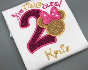 May include: A white t-shirt with pink and gold embroidery. The embroidery includes the number "2", the words "I'm Two-dles!" and the name "Kylie". A pink and gold Minnie Mouse head is above the number.
