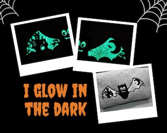 Peut inclure: Un sweat-shirt gris avec un motif de chauve-souris noire dont les ailes présentent une maison hantée et une toile d'araignée. Le texte I GLOW IN THE DARK est écrit en orange. Le motif brille dans le noir avec une lumière cyan vive.