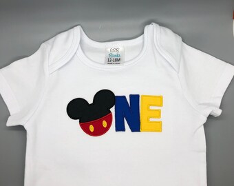 Puede incluir: Mono blanco para bebé con una cabeza de Mickey Mouse y la palabra "ONE" en letras azules, amarillas y rojas. El mono tiene una etiqueta de talla que dice "12-18M".