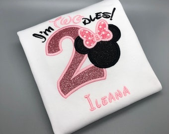 May include: A white t-shirt with pink and black embroidery. The shirt features a pink number "2" with glitter, the words "I'm Two-dles!" and a black and pink Minnie Mouse head. The name "Ileana" is embroidered in pink below the Minnie Mouse head.