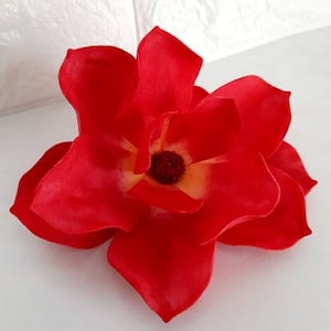 Pode incluir: Uma única flor artificial vermelha grande com um centro amarelo. A flor tem várias pétalas e é feita de um material macio semelhante a tecido.
