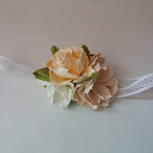 May include: A white lace headband with a cluster of cream and white fabric flowers. The flowers are arranged in a circular shape and have a delicate, feminine look.