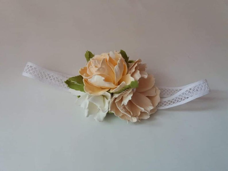 May include: A white lace headband with a cluster of cream and white fabric flowers. The flowers are arranged in a circular shape and have a delicate, feminine look.