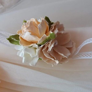 May include: A delicate white headband with a peach and white flower embellishment. The headband is made of lace and features a cluster of fabric flowers.